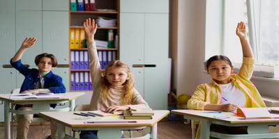 Students raise their hands while sitting at desks in a well-ventilated classroom.
