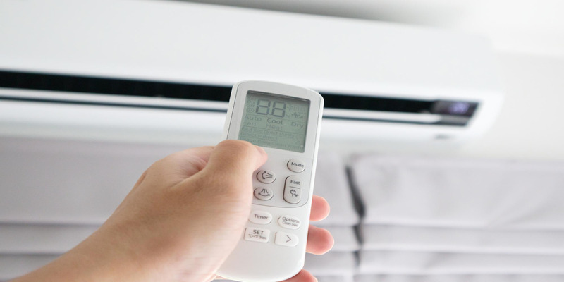 Person holding an air conditioner remote control in front of an AC unit, adjusting the settings.