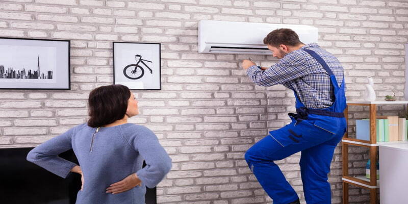 Technician servicing an air conditioner while a woman observes in the background.