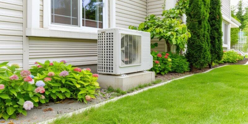 The outdoor unit of a mini-split air conditioner is installed near the house on a concrete stand among greenery.
