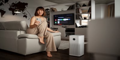 Woman sitting in a modern living room with a smartphone, next to an air purifier running on the floor.